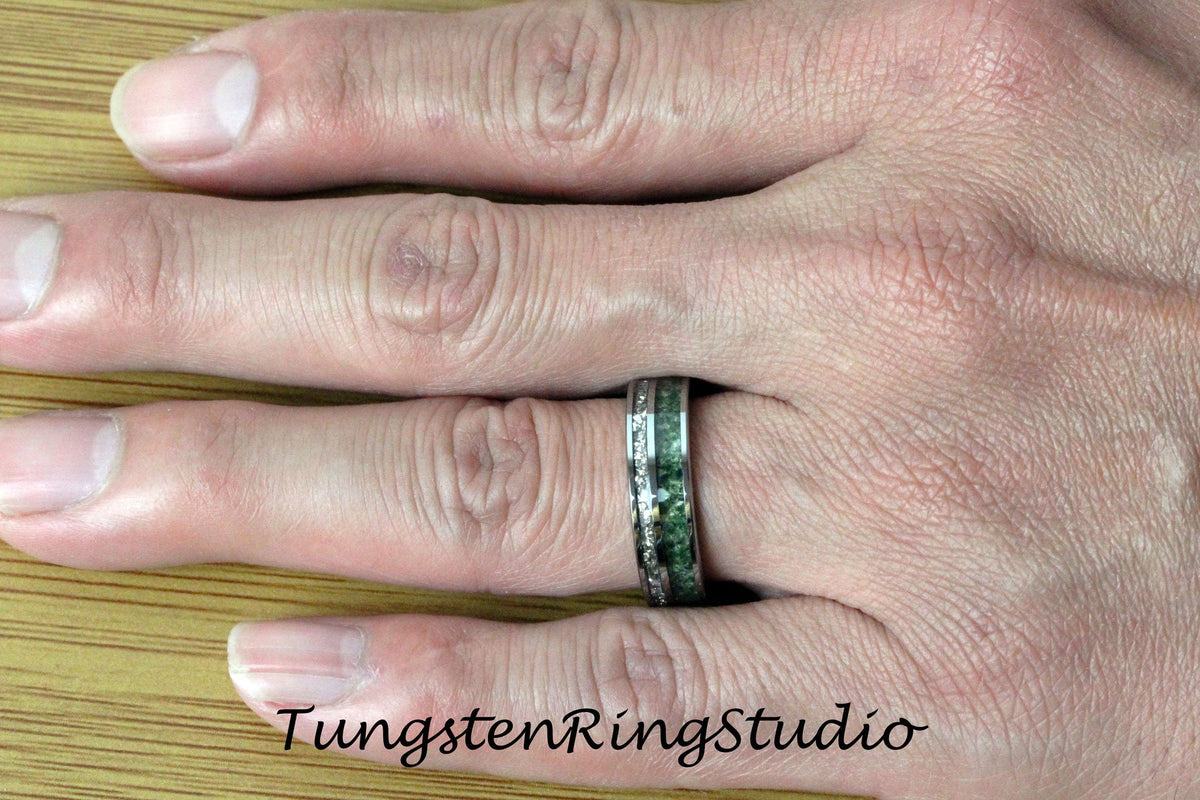 May include: A silver tungsten ring with a green and silver inlay. The ring is on a person's finger.