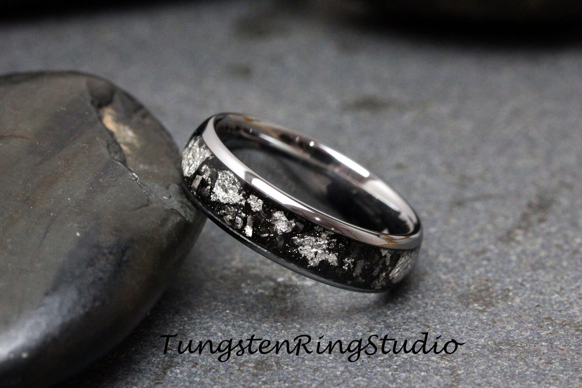 May include: A silver tungsten ring with a black inlay and silver flecks. The ring is on a gray stone surface.