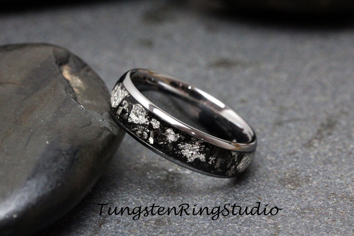 May include: A silver tungsten ring with a black inlay and silver flecks. The ring is on a gray stone surface.