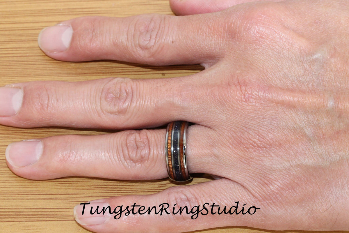 May include: A tungsten ring with a black and brown wood inlay. The ring is on a person's finger.