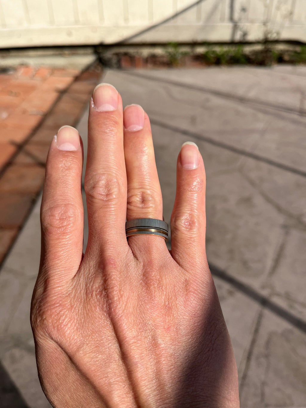 May include: A silver and gold wedding band with a brushed finish. The band is worn on the ring finger of a person's left hand.