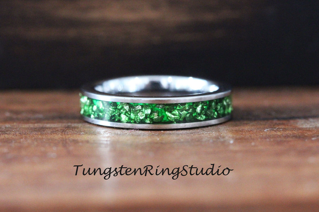 May include: A silver tungsten ring with a green crushed glass inlay. The ring is on a wooden surface. TungstenRingStudio.