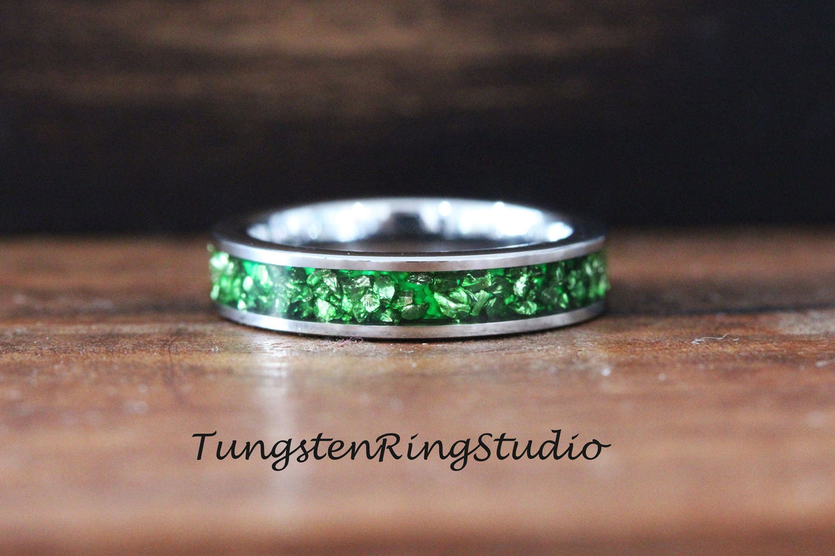 May include: A silver tungsten ring with a green crushed glass inlay. The ring is on a wooden surface. TungstenRingStudio.