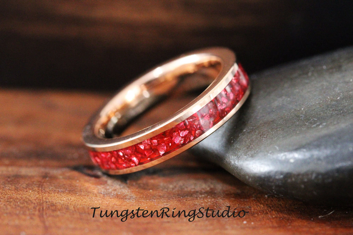 May include: A rose gold tungsten carbide ring with a red inlay of crushed gemstones. The ring is a simple band with a smooth finish.