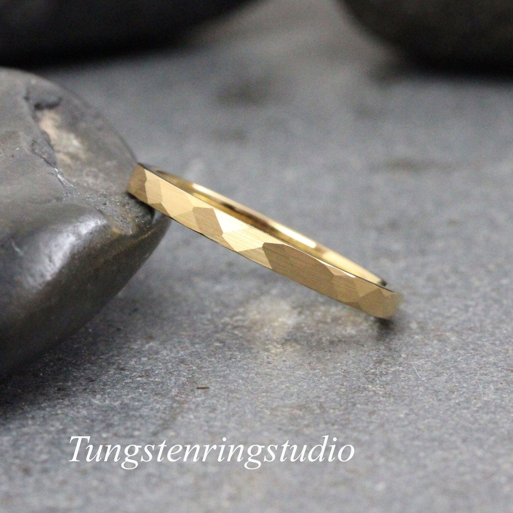 May include: A gold hammered wedding band with a textured finish. The band is thin and delicate, and it is shown on a gray stone background.