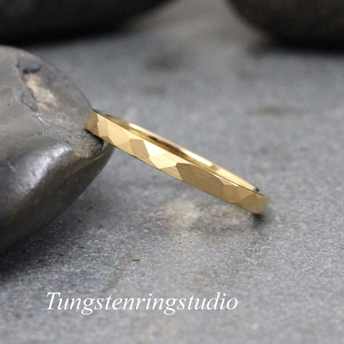 May include: A gold hammered wedding band with a textured finish. The band is thin and delicate, and it is shown on a gray stone background.