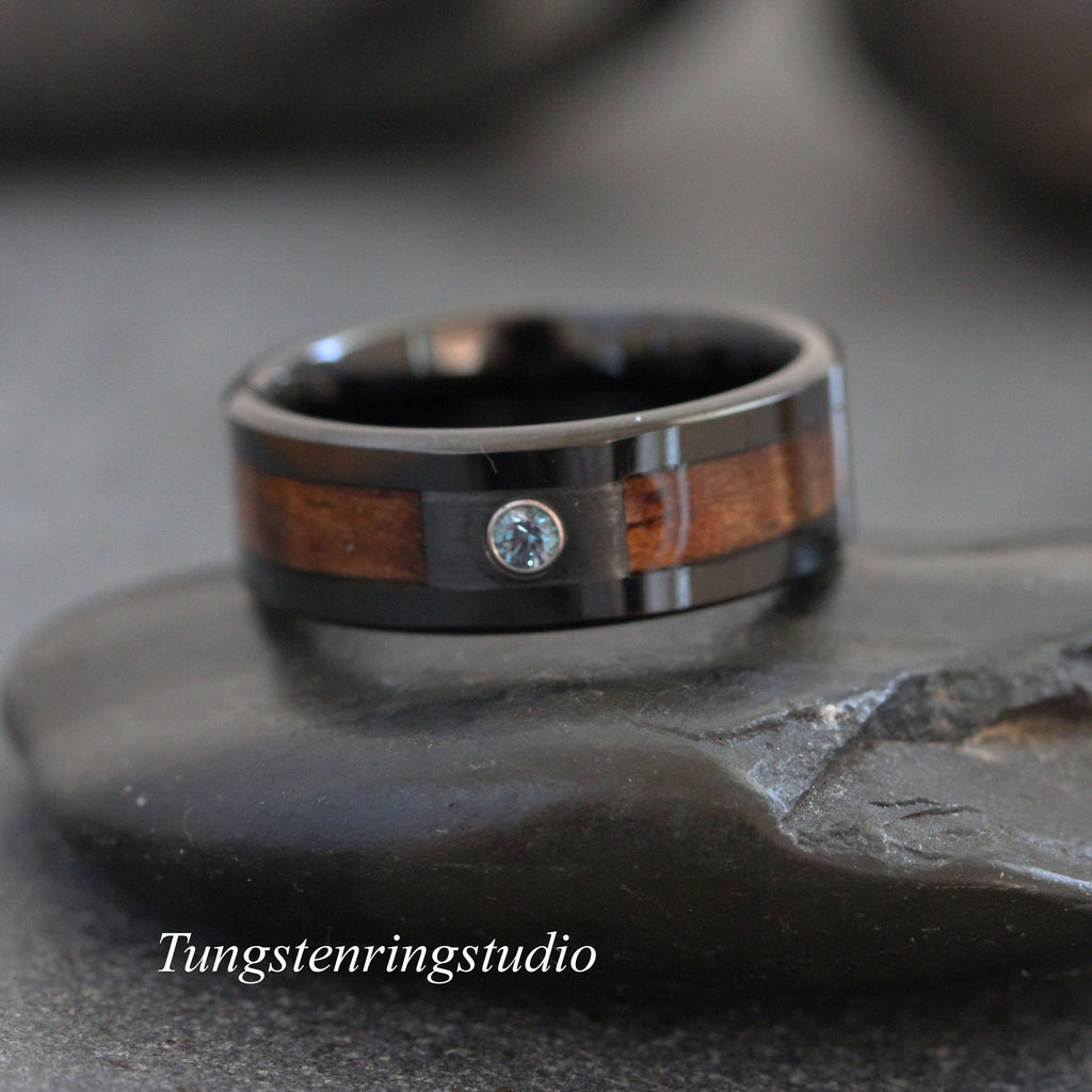 May include: A black ring featuring a central inlay of brown wood and a single small, round, light blue gemstone set in the metal. The text Tungstenringstudio is visible in white lettering at the bottom left of the image frame.