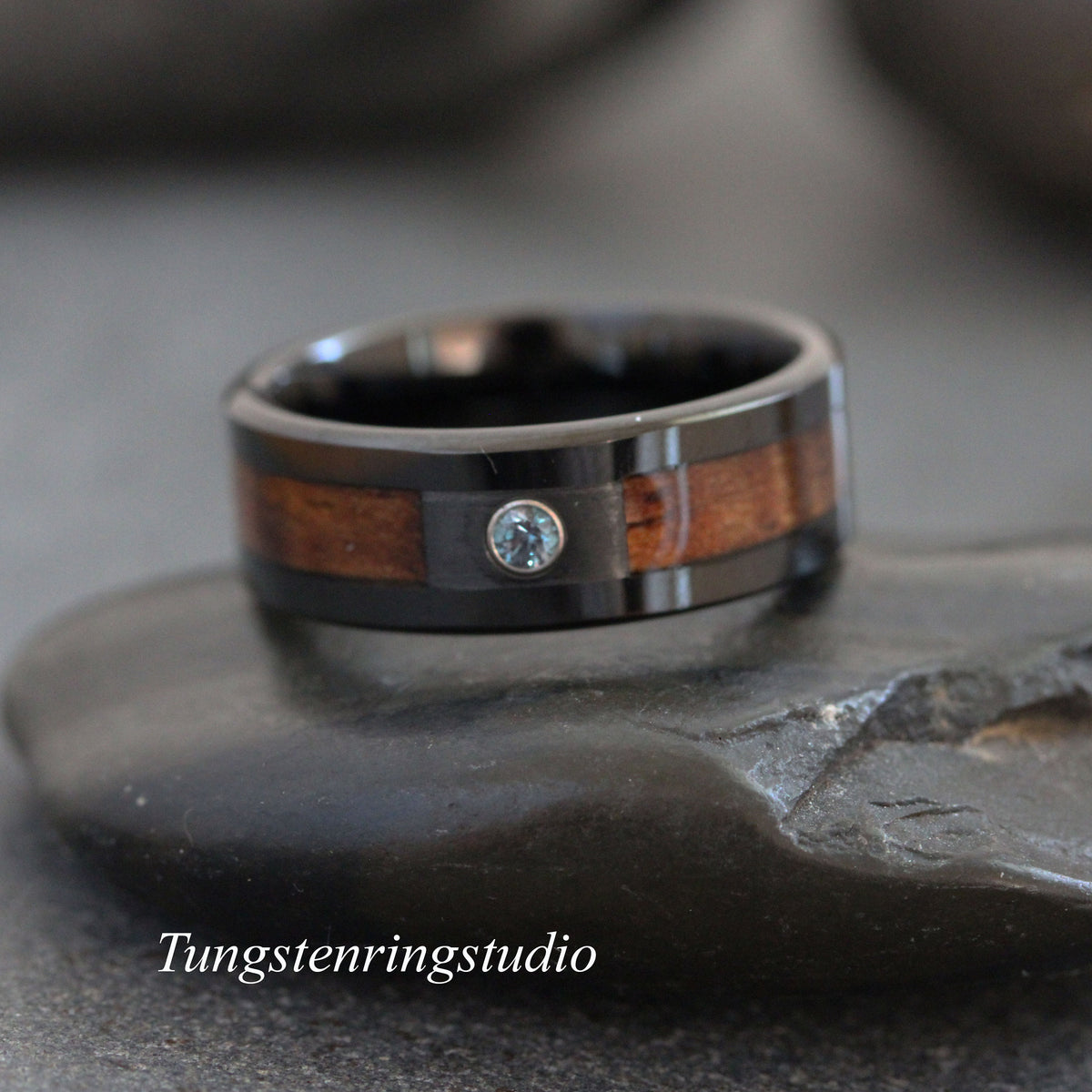 May include: A black ring featuring a central inlay of brown wood and a single small, round, light blue gemstone set in the metal. The text Tungstenringstudio is visible in white lettering at the bottom left of the image frame.
