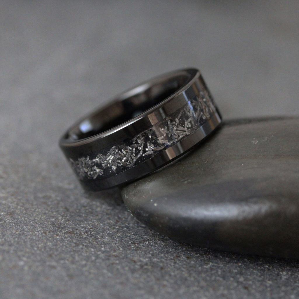 May include: A wide, polished black band ring featuring a central inlay of textured, metallic silver-toned material with an irregular, fragmented pattern that wraps around the circumference of the ring, creating a distinct contrast against the dark metal.