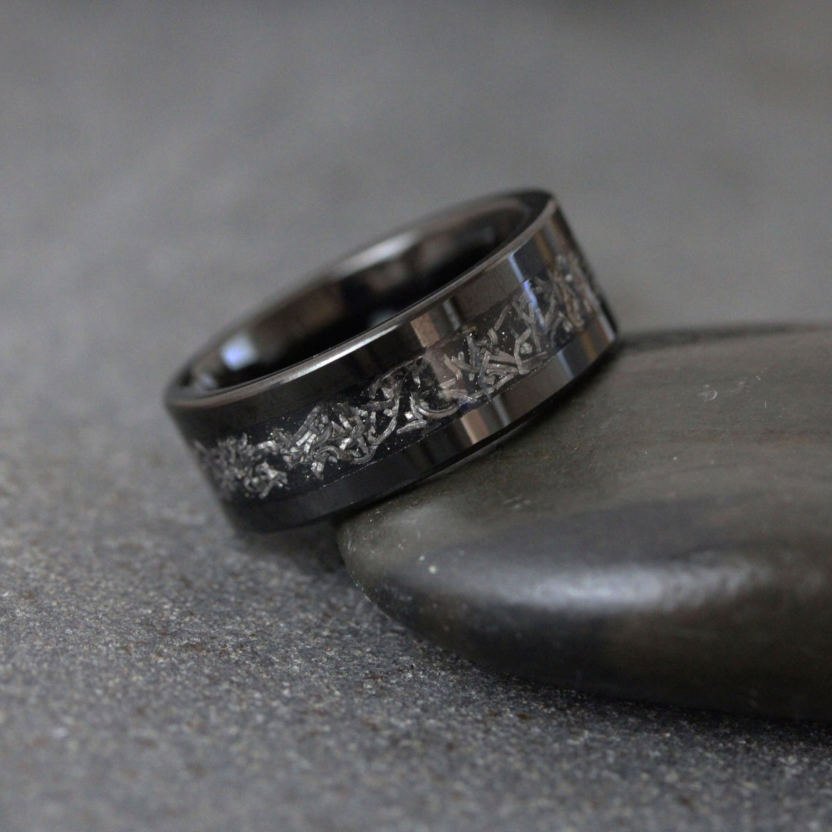 May include: A wide, polished black band ring featuring a central inlay of textured, metallic silver-toned material with an irregular, fragmented pattern that wraps around the circumference of the ring, creating a distinct contrast against the dark metal.