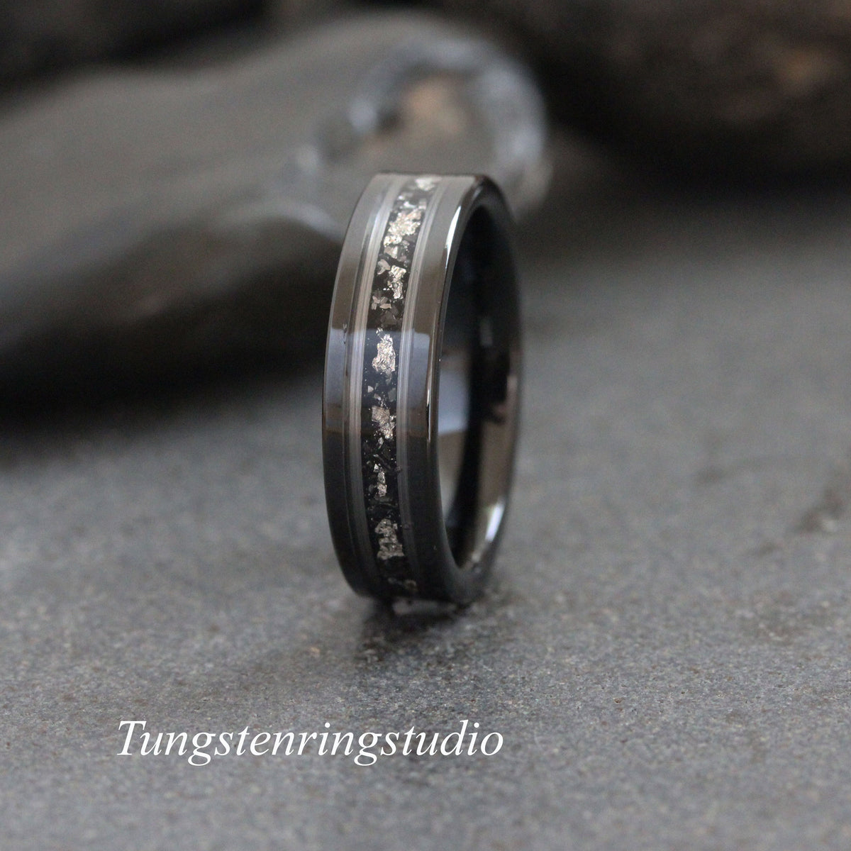 May include: A black tungsten carbide ring with a silver inlay. The inlay is a thin line of silver that runs around the ring. The ring is polished and has a smooth finish.