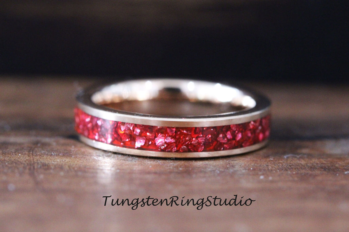May include: A silver tungsten ring with a red inlay of crushed gemstones. The ring is on a wooden surface. TungstenRingStudio.