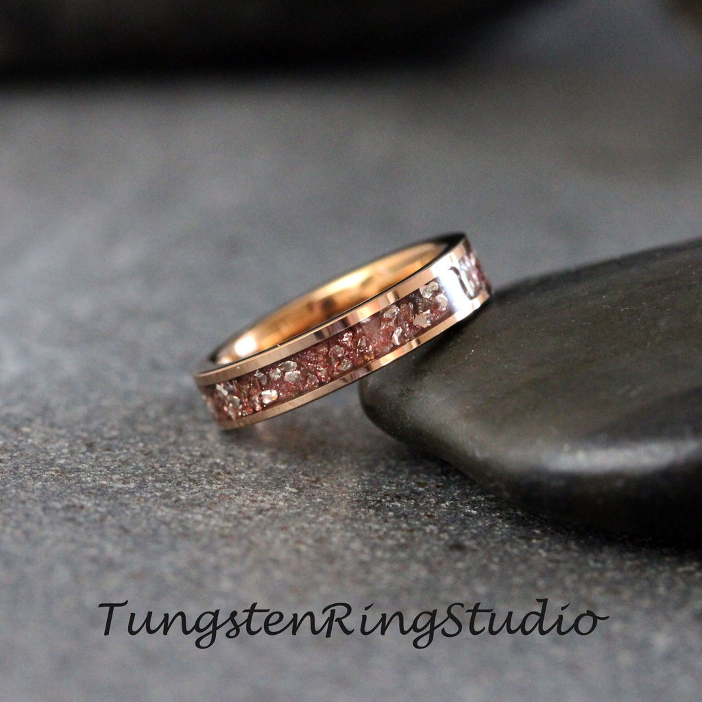 May include: A rose gold tungsten carbide ring with a band of embedded pink and white particles. The ring is on a gray stone surface.