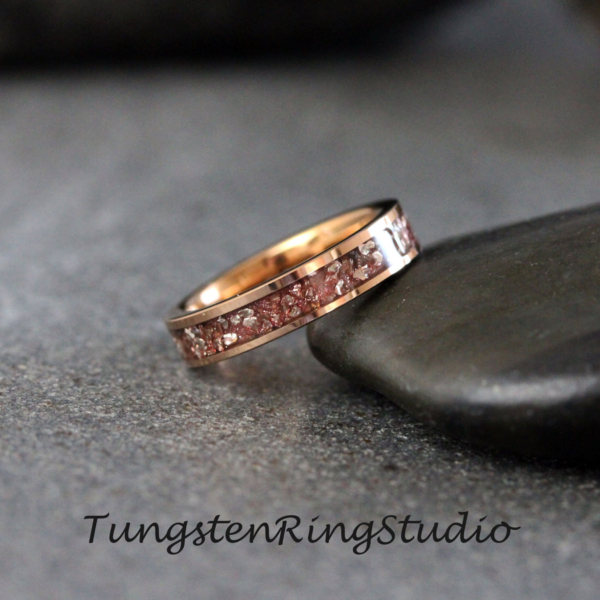 May include: A rose gold tungsten carbide ring with a band of embedded pink and white particles. The ring is on a gray stone surface.