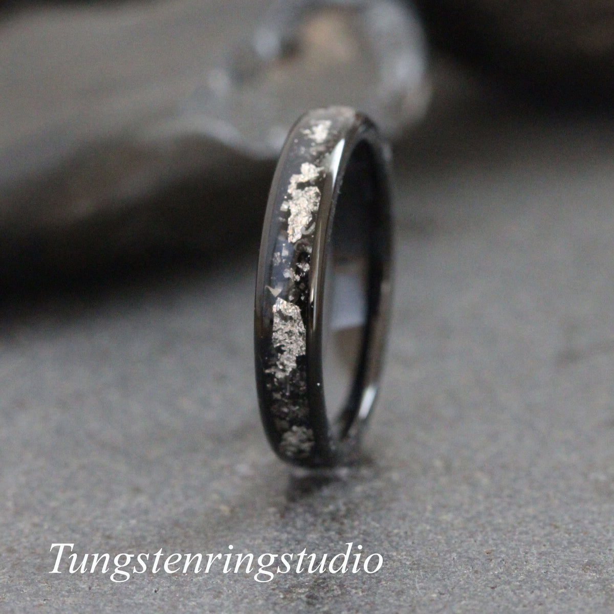 May include: A black tungsten carbide ring with a silver inlay. The inlay is in a scattered pattern. Tungstenringsstudio is printed below the ring.
