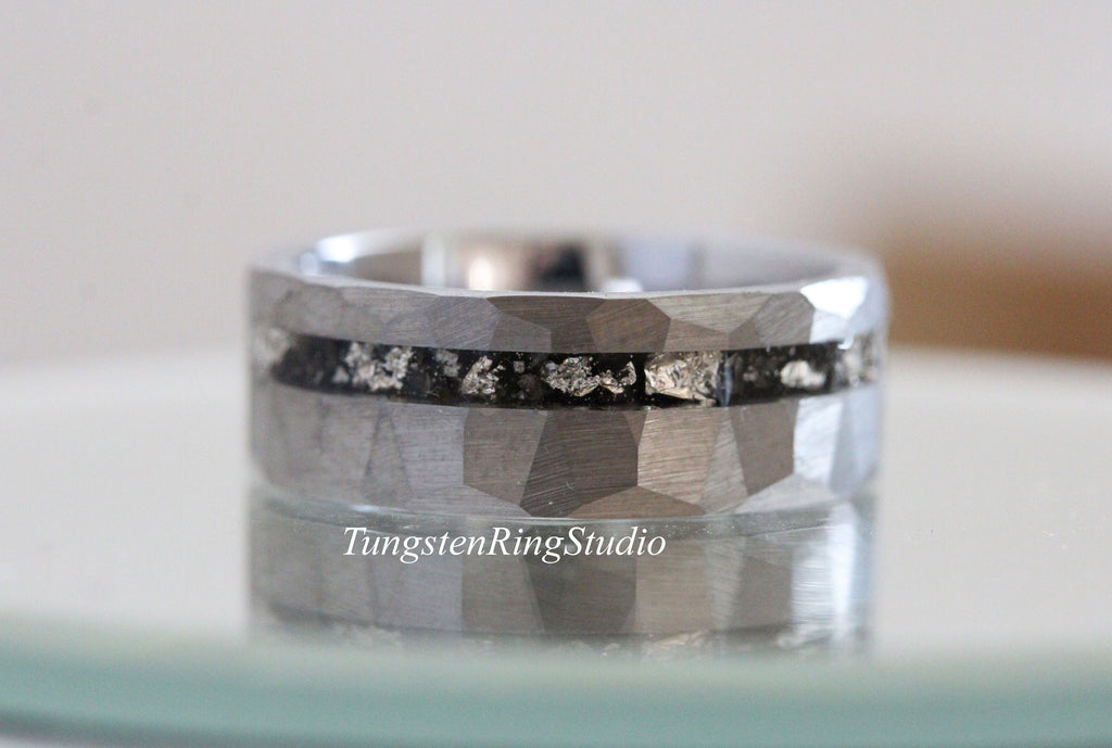 May include: A silver tungsten ring with a hammered finish and a black inlay with silver flecks. The ring is shown on a reflective surface.
