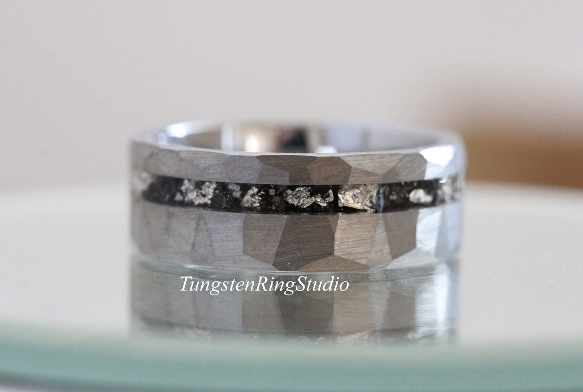 May include: A silver tungsten ring with a hammered finish and a black inlay with silver flecks. The ring is shown on a reflective surface.