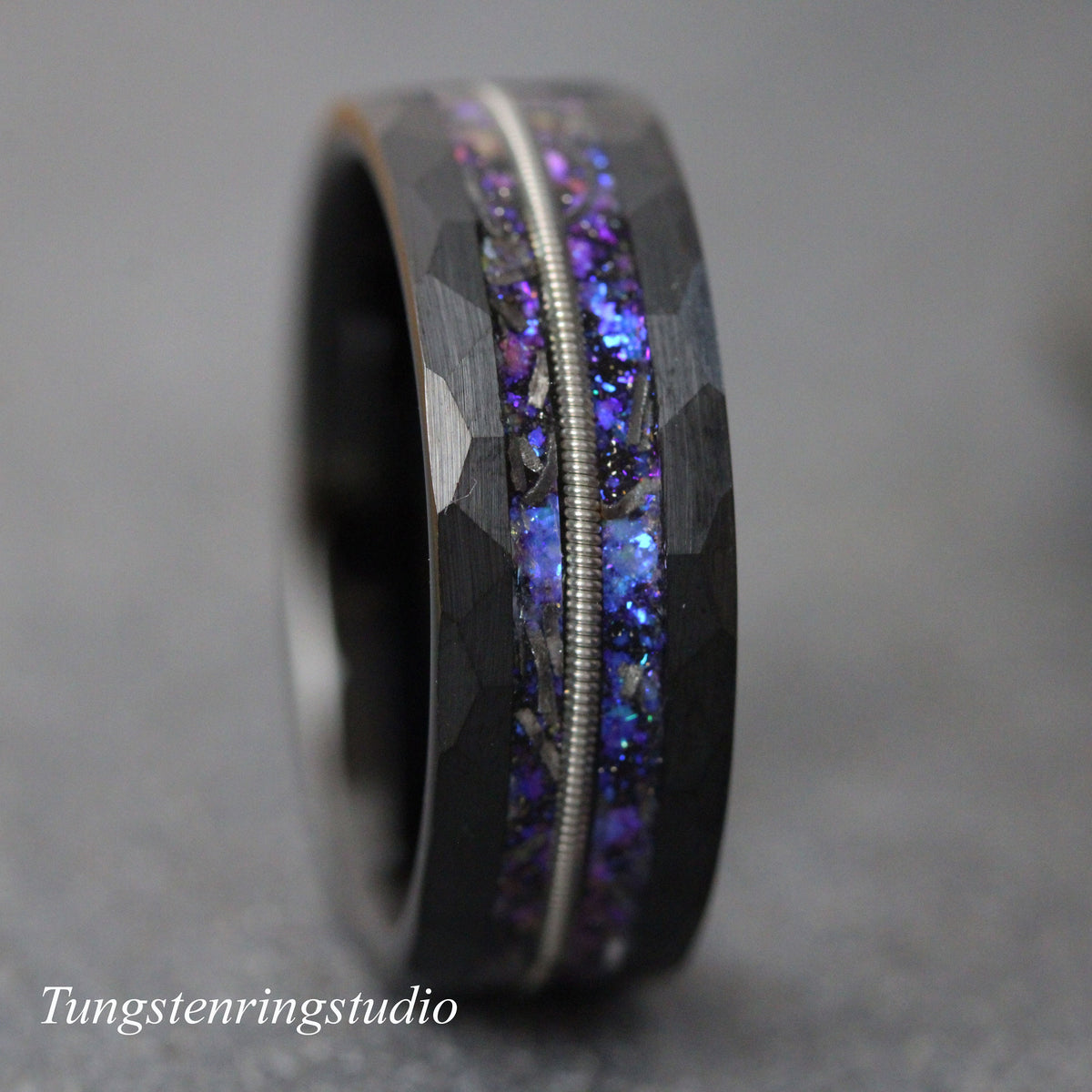 May include: A black tungsten carbide ring with a purple and silver inlay. The ring has a hammered finish.