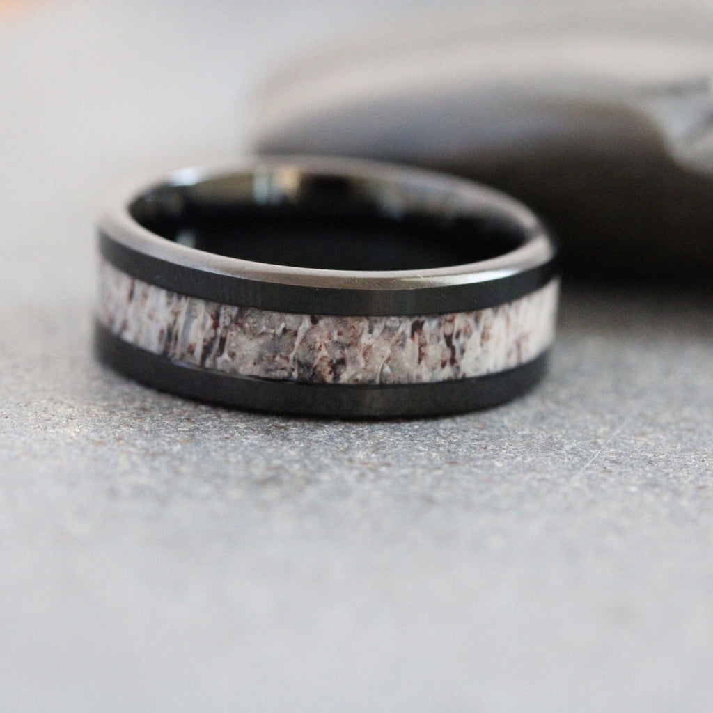 May include: A black metal ring featuring a central inlay of textured, light-colored organic material with brown and grey mottled patterns. The band has a smooth, polished finish on the top and bottom edges, framing the rough-textured inlay.