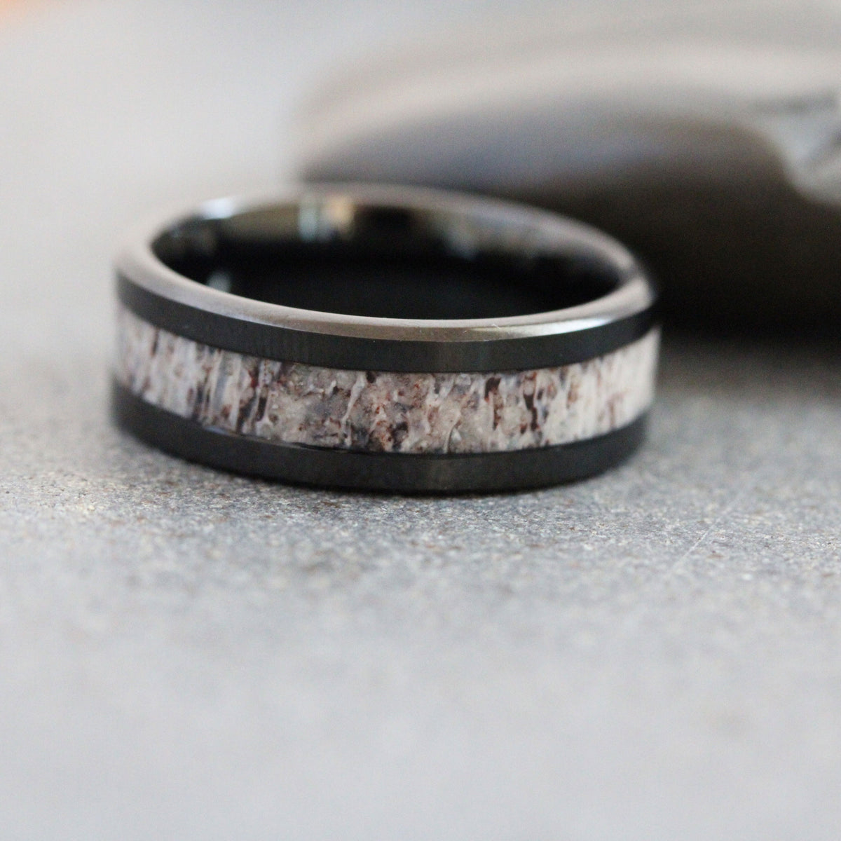May include: A black metal ring featuring a central inlay of textured, light-colored organic material with brown and grey mottled patterns. The band has a smooth, polished finish on the top and bottom edges, framing the rough-textured inlay.