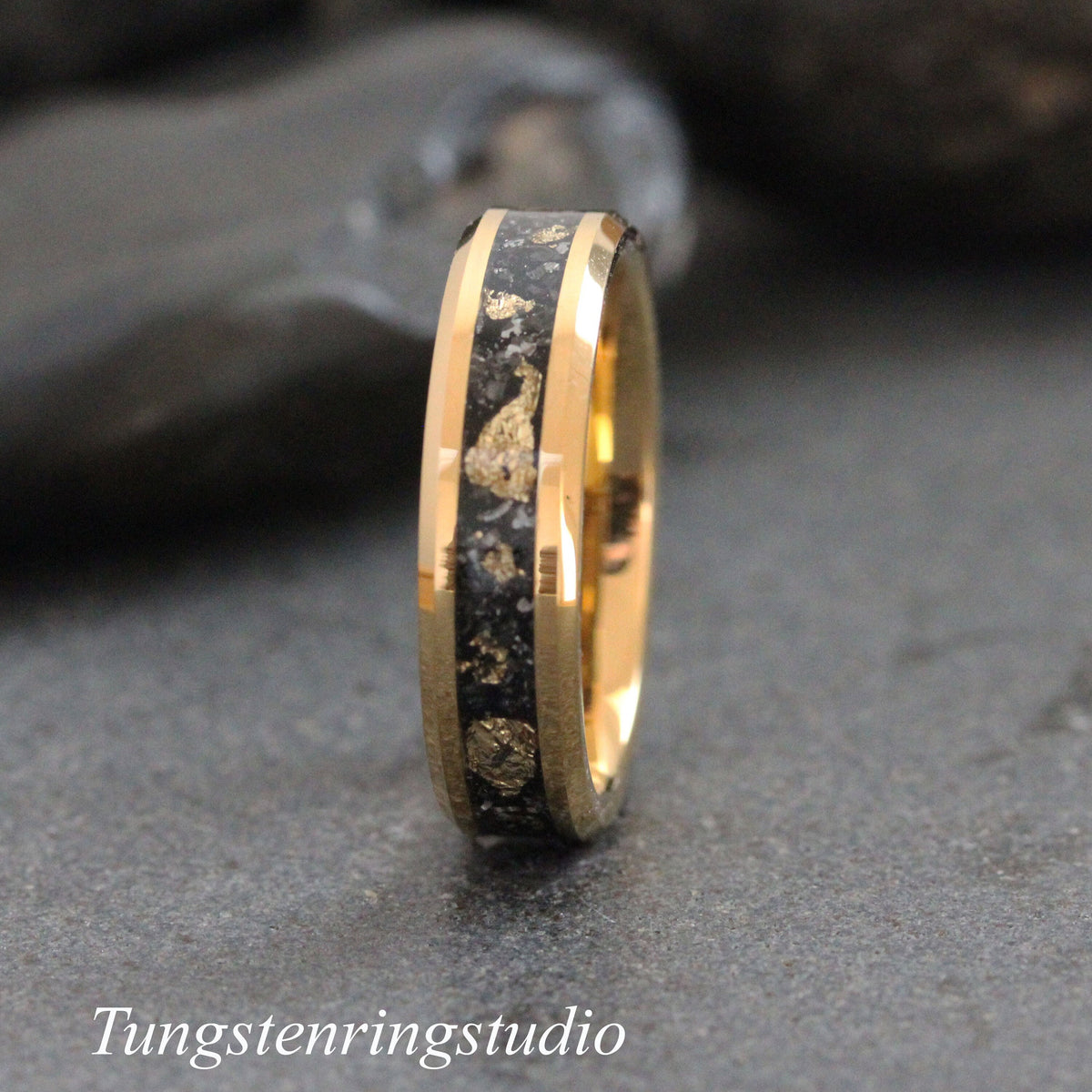 May include: A gold-colored ring with a black inlay speckled with gold-colored flakes. The ring has a polished finish and the text "Tungstenringstudio" is at the bottom of the image. This ring is a piece of jewelry.