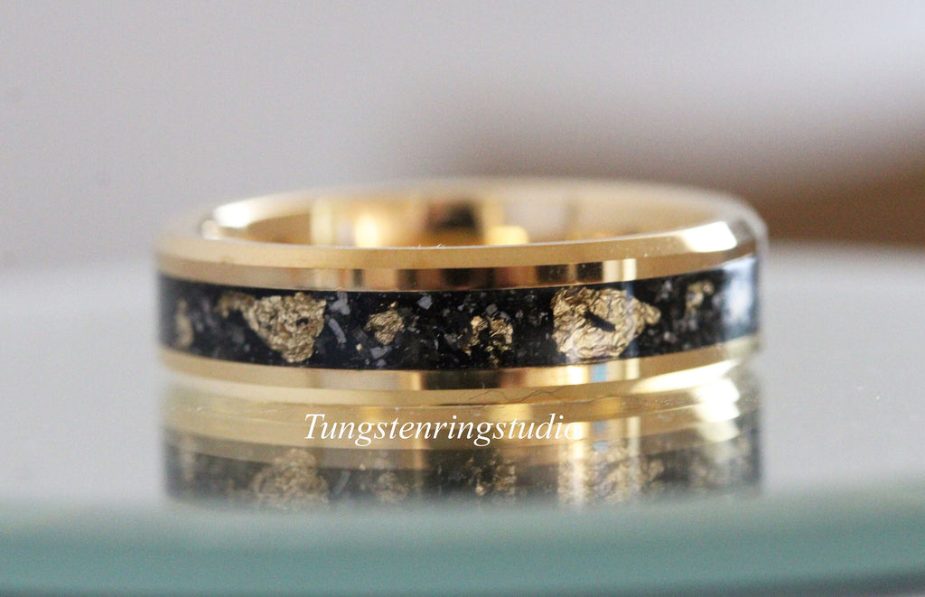 May include: A gold-colored ring with a black inlay speckled with gold flakes. The ring has a polished finish and the text "Tungstenringstudio" is visible on the ring. This ring is a piece of jewelry.