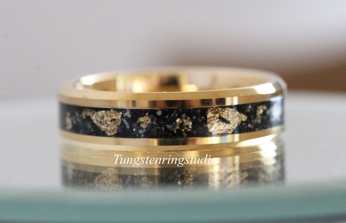 May include: A gold-colored ring with a black inlay speckled with gold flakes. The ring has a polished finish and the text "Tungstenringstudio" is visible on the ring. This ring is a piece of jewelry.