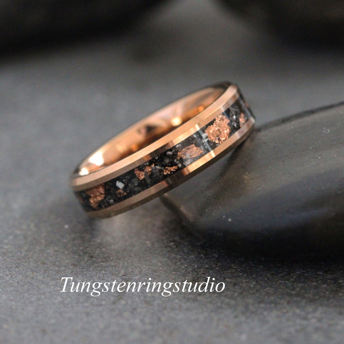 May include: A rose gold tungsten carbide ring with a black inlay and copper flecks. The ring is a classic style with a beveled edge.