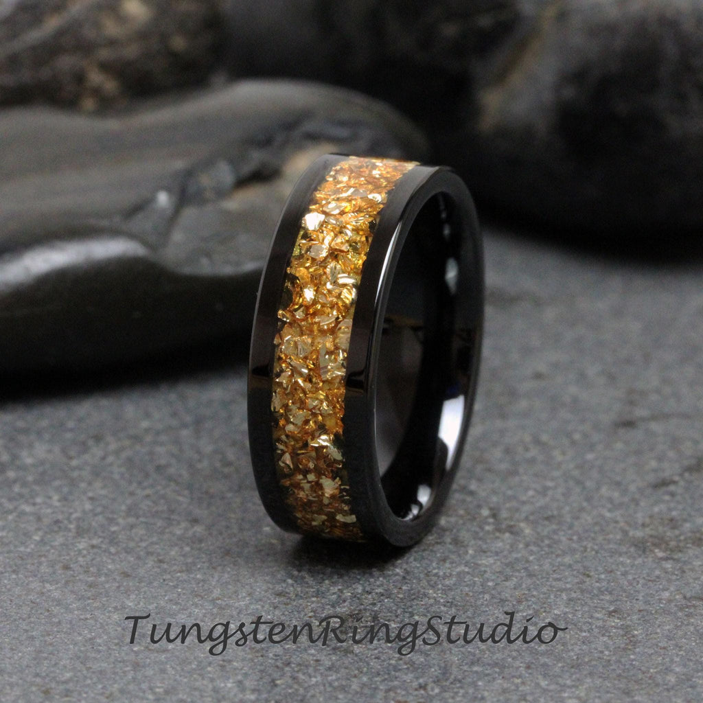 May include: A black tungsten carbide ring with a gold inlay of small, irregular pieces. The ring is polished and has a smooth finish.