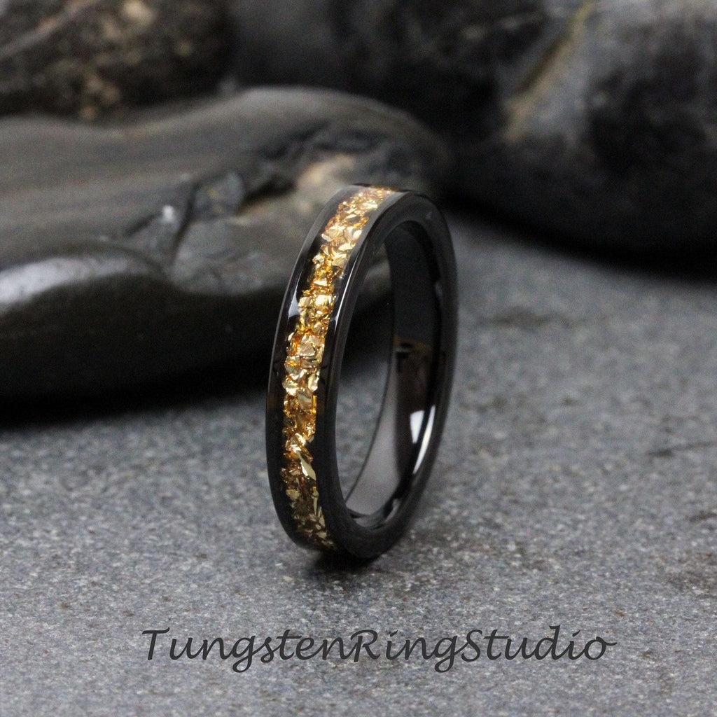 May include: A black tungsten carbide ring with a band of gold flakes. Tungsten Ring Studio is printed on the surface below the ring.