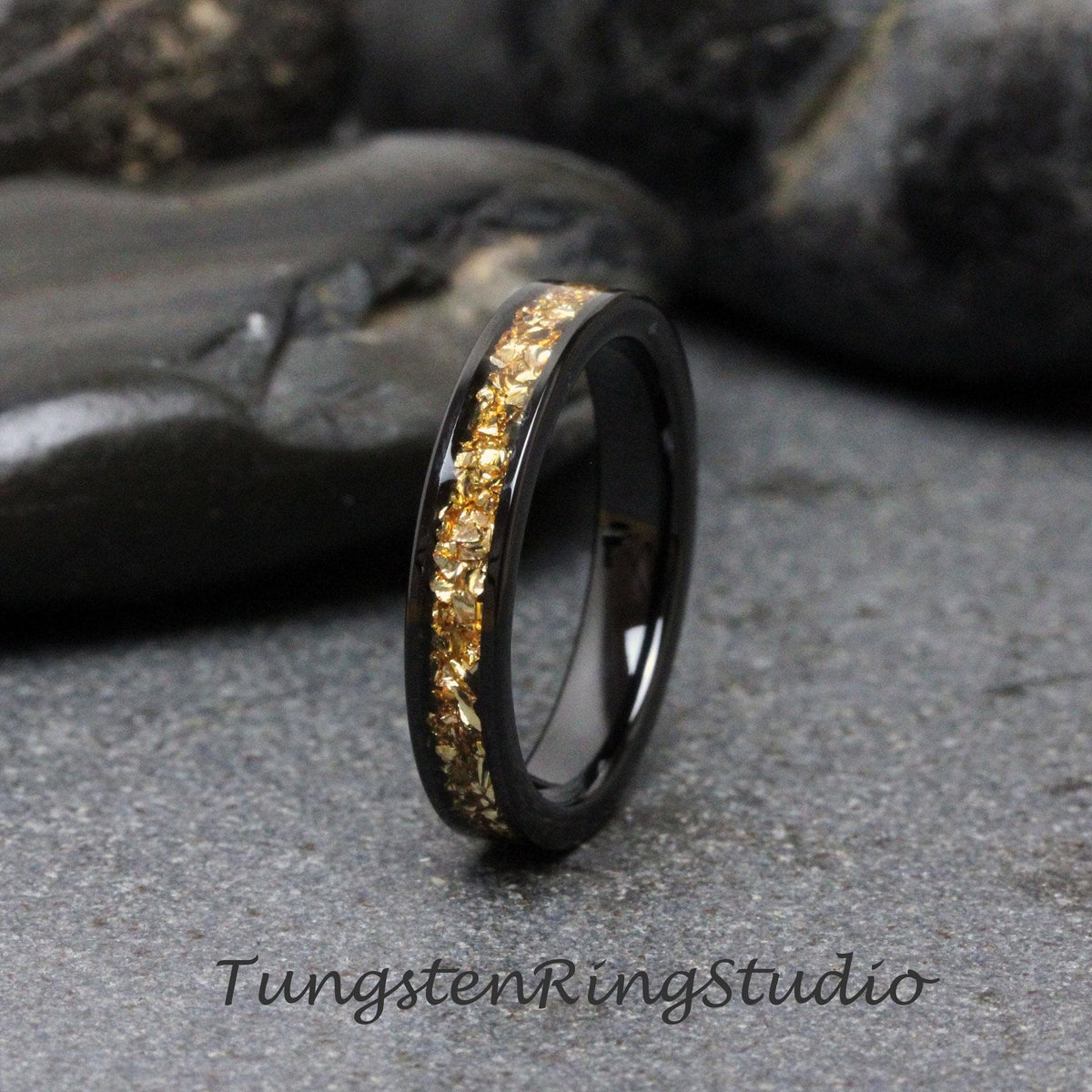 May include: A black tungsten carbide ring with a band of gold flakes. Tungsten Ring Studio is printed on the surface below the ring.