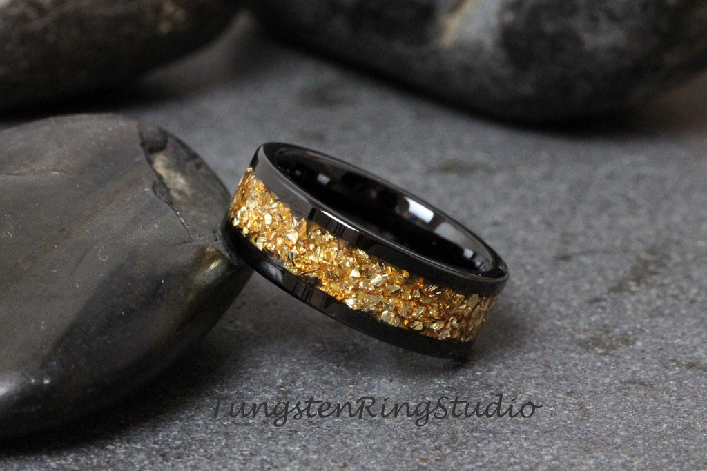 May include: A black tungsten carbide ring with a gold inlay of small, irregular pieces. The ring is a simple band with a smooth, polished finish.