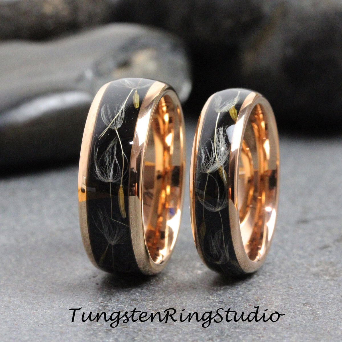 May include: Two matching wedding bands with a rose gold finish. The bands feature a black inlay with delicate white dandelion seeds.