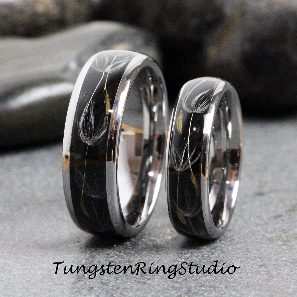 May include: Two black tungsten carbide wedding bands with a silver inlay featuring a dandelion design. The bands are polished and have a beveled edge.