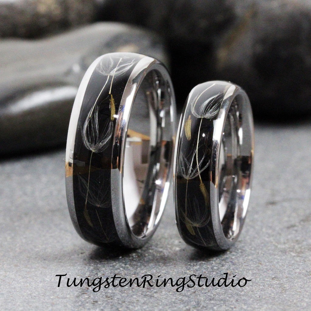 May include: Two black tungsten carbide wedding bands with a silver inlay featuring a dandelion design. The rings are shown side-by-side.