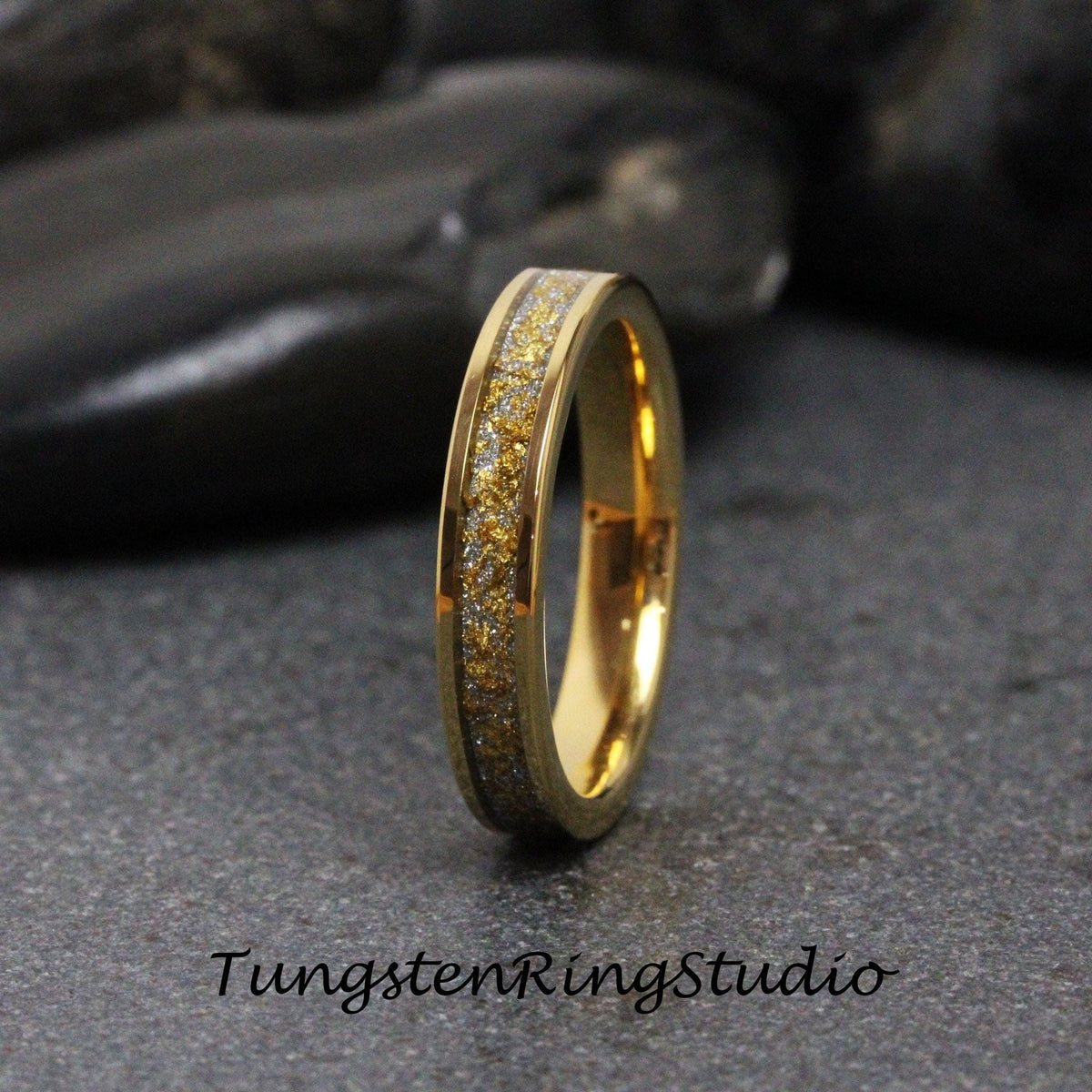 May include: A gold-colored ring with a band of gold and silver flecks. The ring has a polished finish and the text "TungstenRingStudio" is at the bottom. The ring is on a dark gray surface.