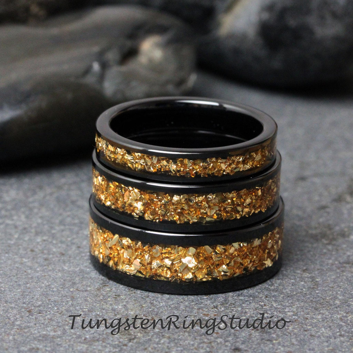 May include: Stack of four black tungsten carbide rings with a gold inlay. The rings are stacked on a gray surface.