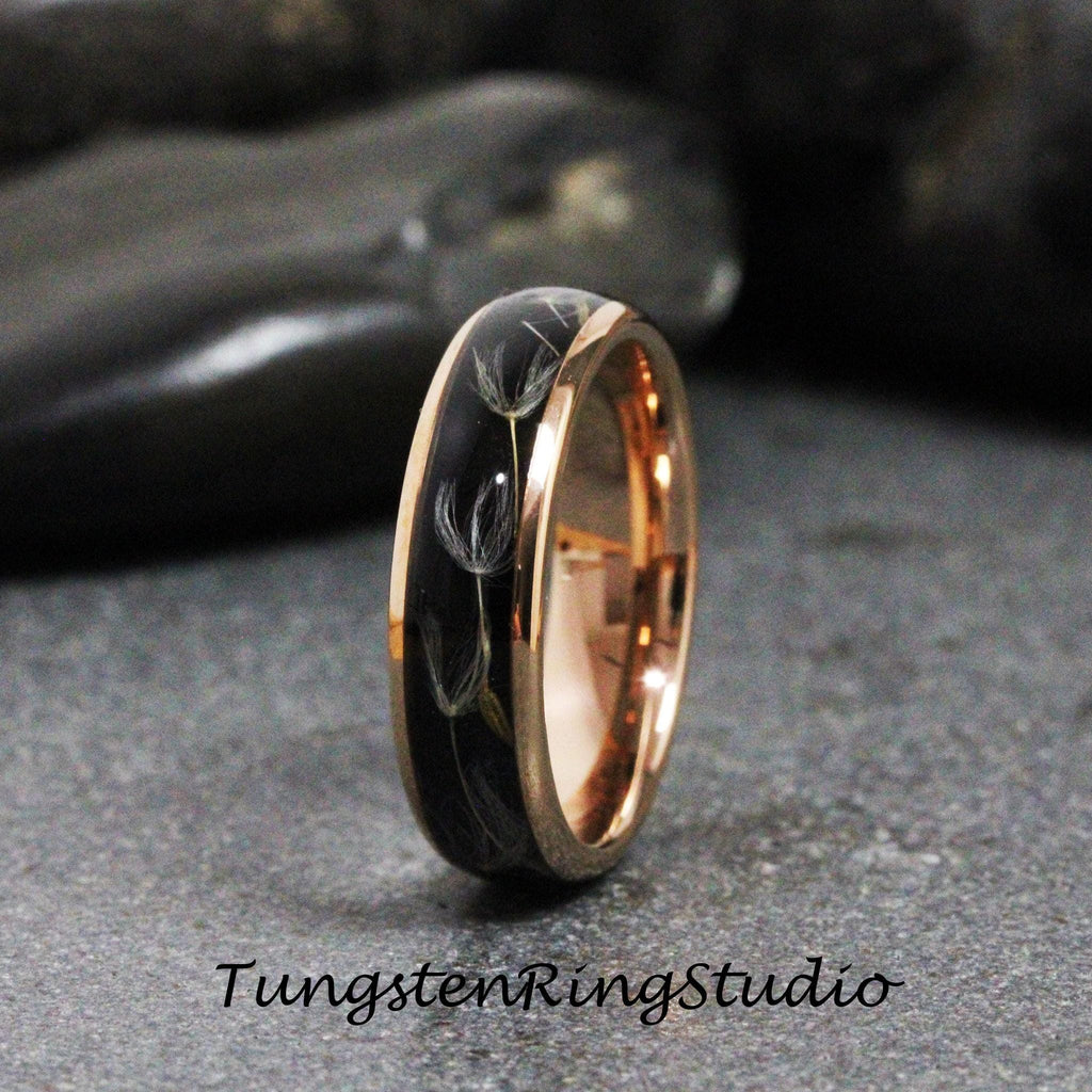 May include: A black tungsten ring with a rose gold band and a design of dandelion seeds embedded in the black band. The ring is on a dark gray surface.