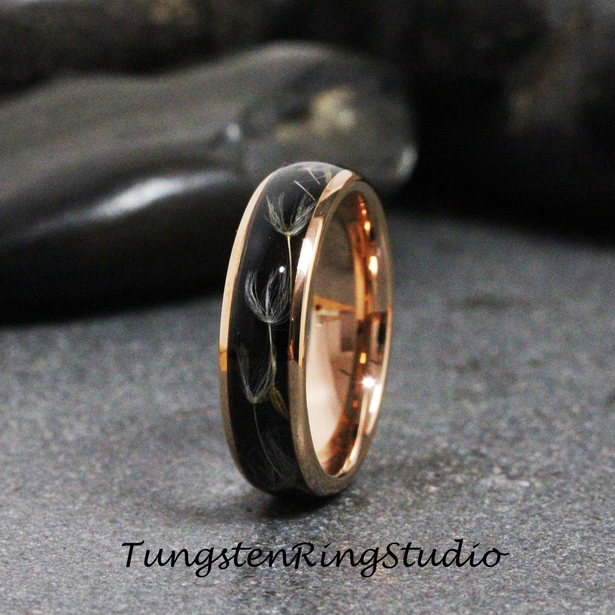 May include: A black tungsten ring with a rose gold band and a design of dandelion seeds embedded in the black band. The ring is on a dark gray surface.