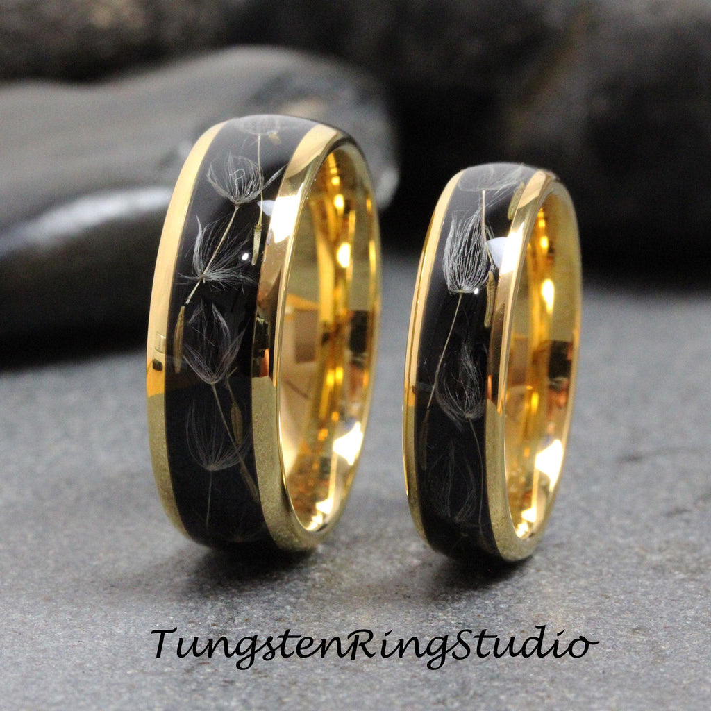 May include: Two tungsten carbide wedding bands with a black inlay and gold trim. The inlay features a design of dandelion seeds.