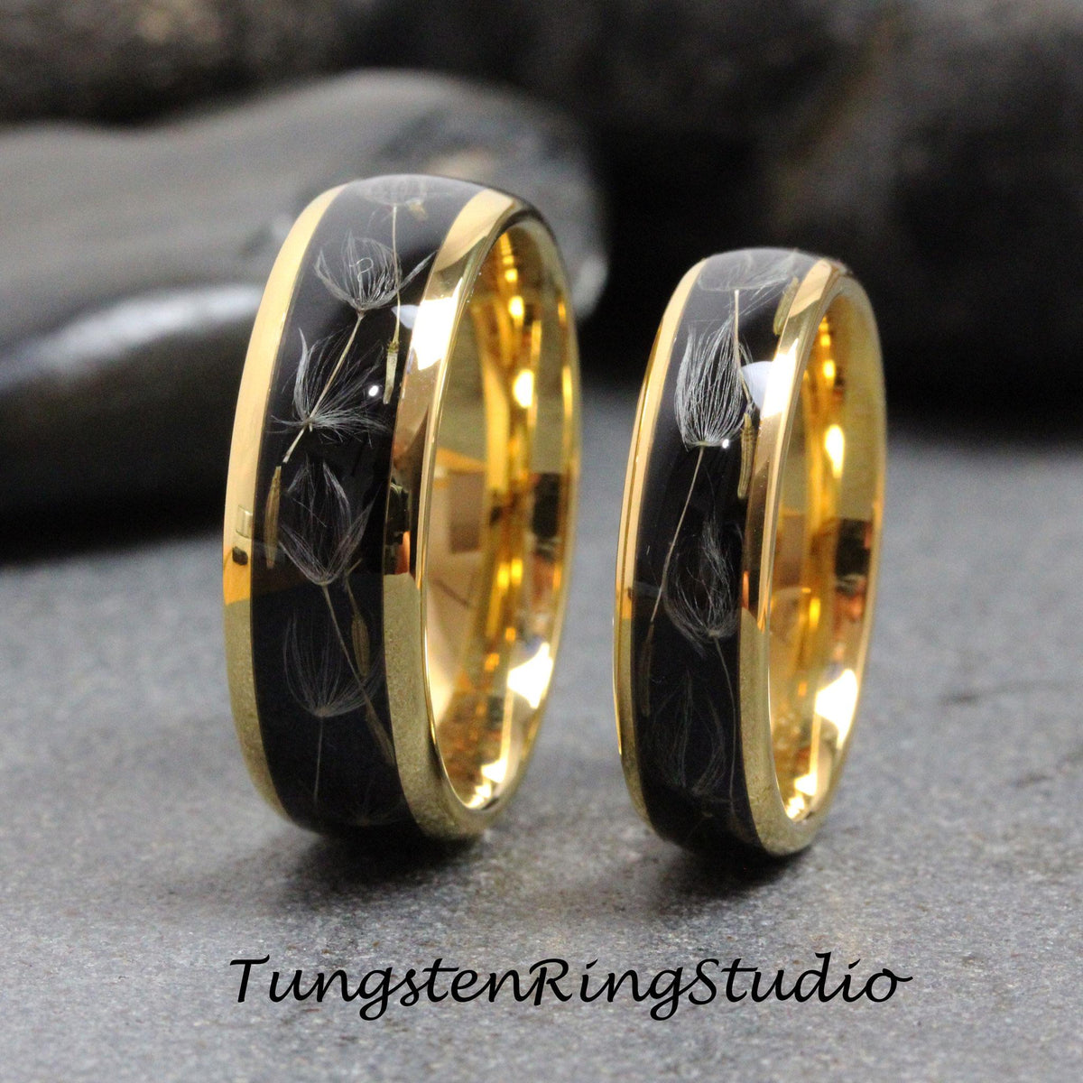 May include: Two tungsten carbide wedding bands with a black inlay and gold trim. The inlay features a design of dandelion seeds.