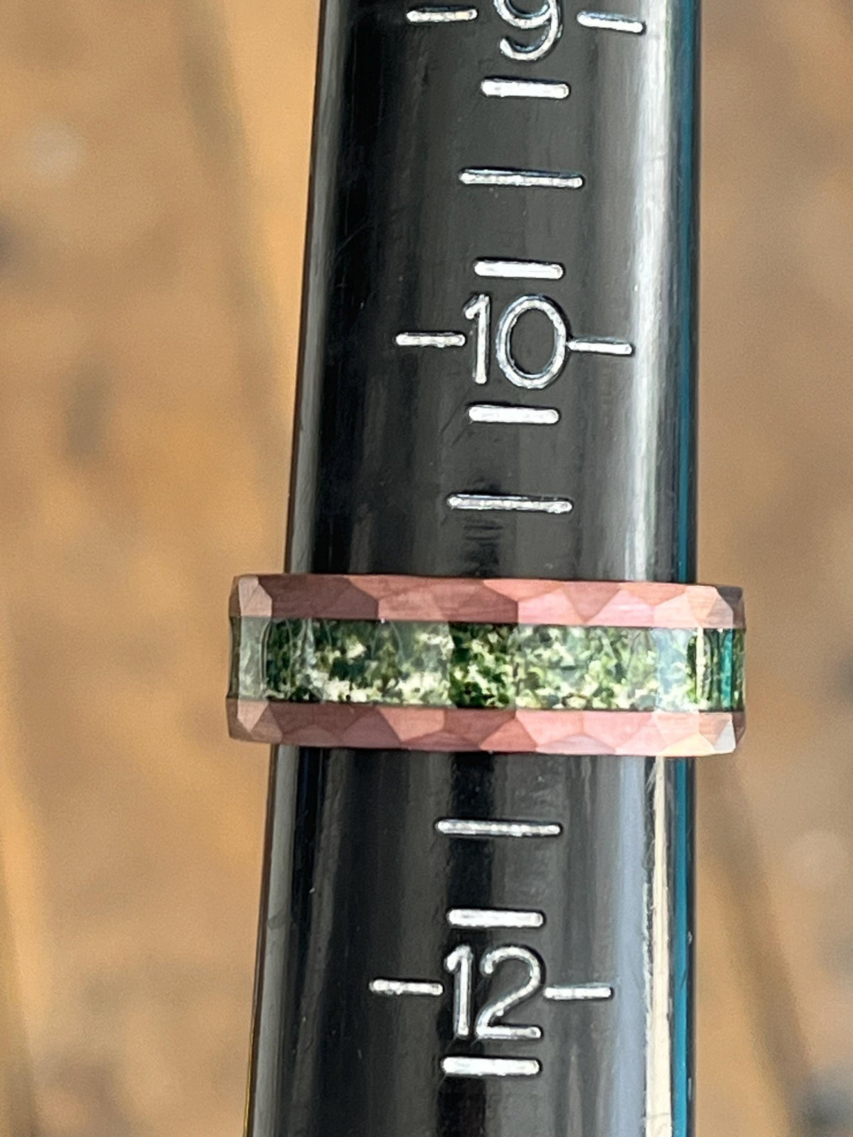 May include: A close-up of a dark green and pink ring with a textured surface. The ring is on a black ruler with white markings. The ruler shows the numbers 9, 10, and 12.