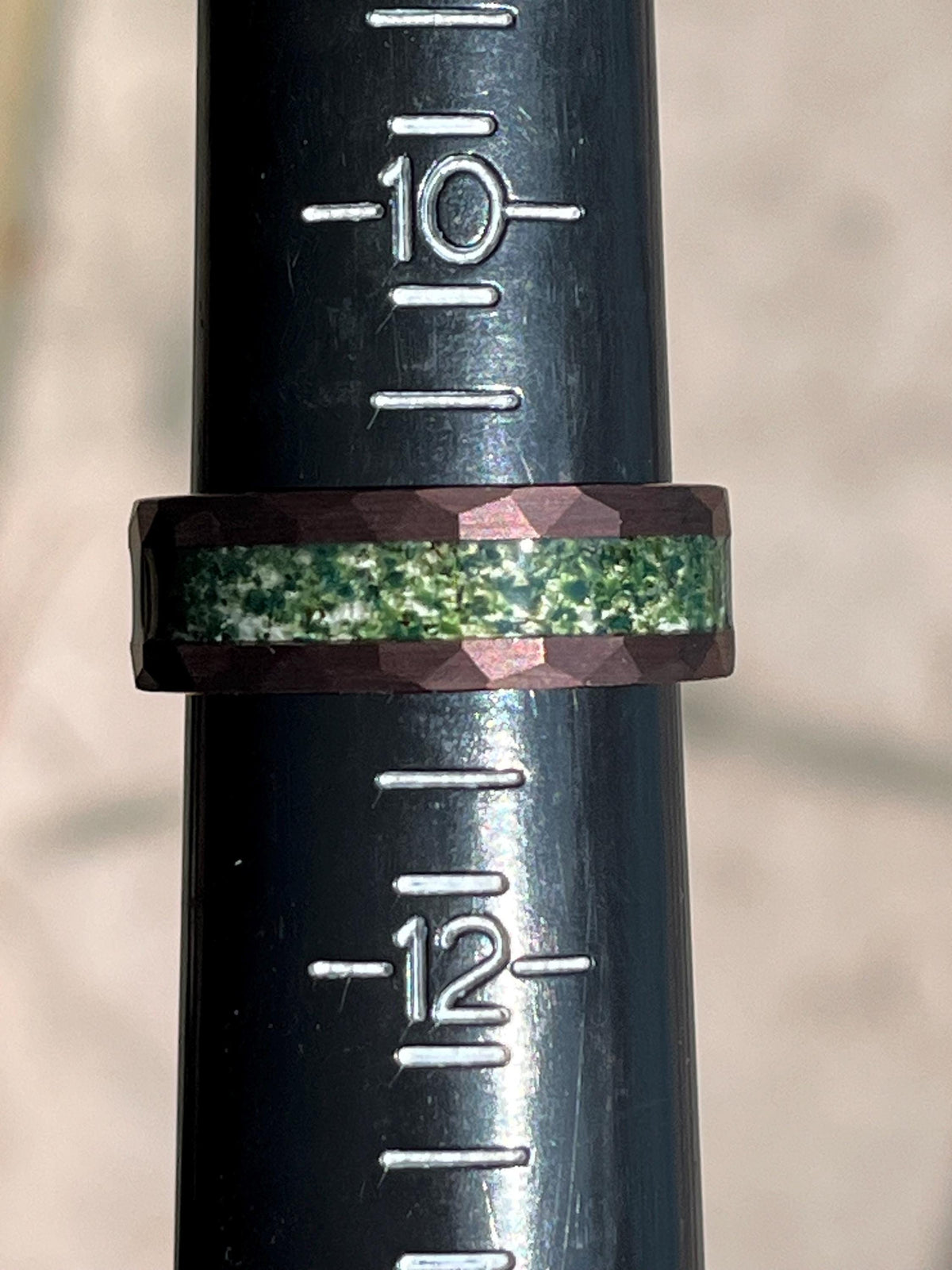 May include: A dark brown and green ring with a textured band. The ring is measured against a ruler marked with numbers 10 and 12.