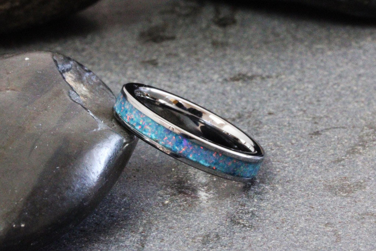 May include: A silver ring with a band of blue and pink opal-like material. The ring is set against a dark gray stone and a textured gray surface. The ring's design is simple and elegant.