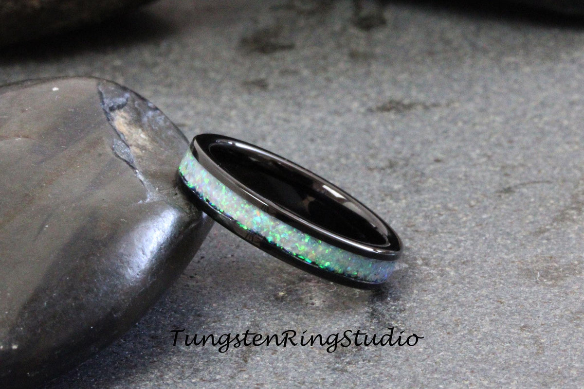 May include: A black tungsten ring with a white opal inlay. The ring is on a gray stone surface. TungstenRingStudio is printed on the surface.