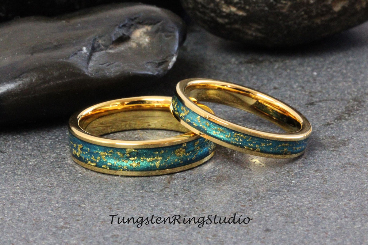 May include: Two gold wedding bands with a teal blue and gold inlay. The bands are on a dark gray background.