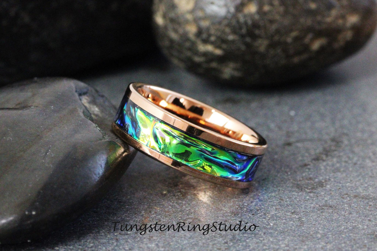May include: A rose gold tungsten carbide ring with a green, blue, and iridescent abalone shell inlay. The ring is on a dark gray background.