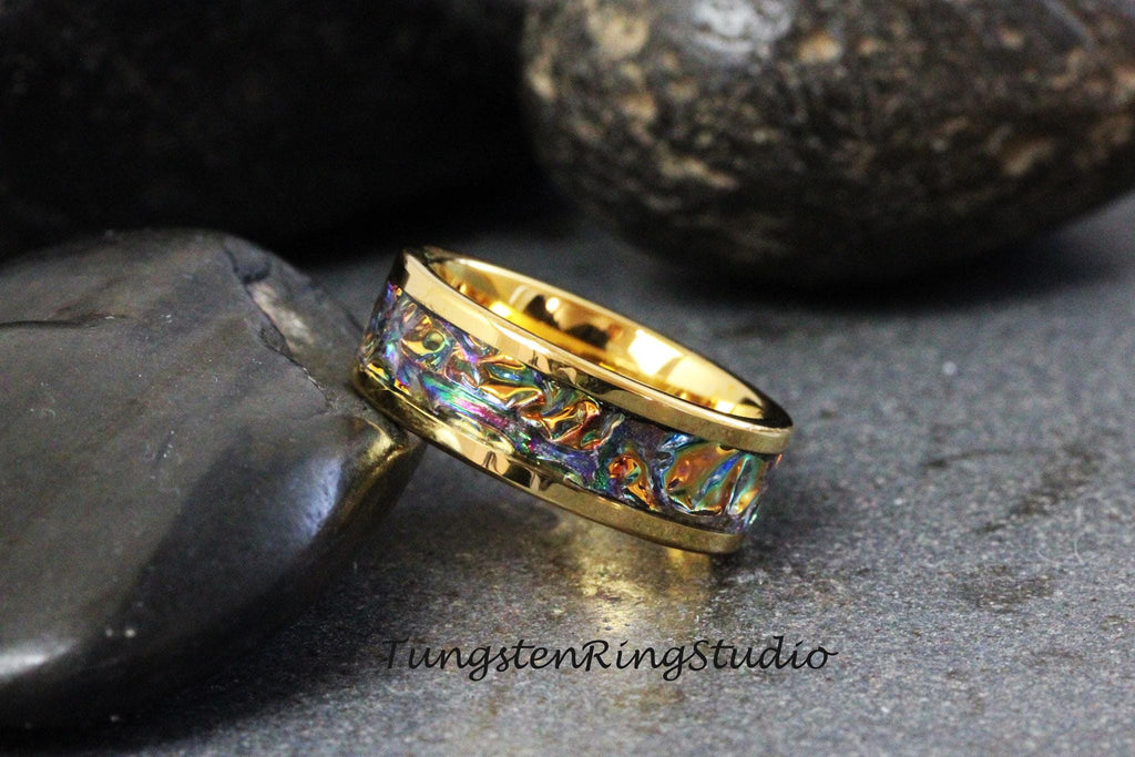 May include: A gold tungsten carbide ring with a colorful, iridescent inlay. The ring is on a dark gray background.