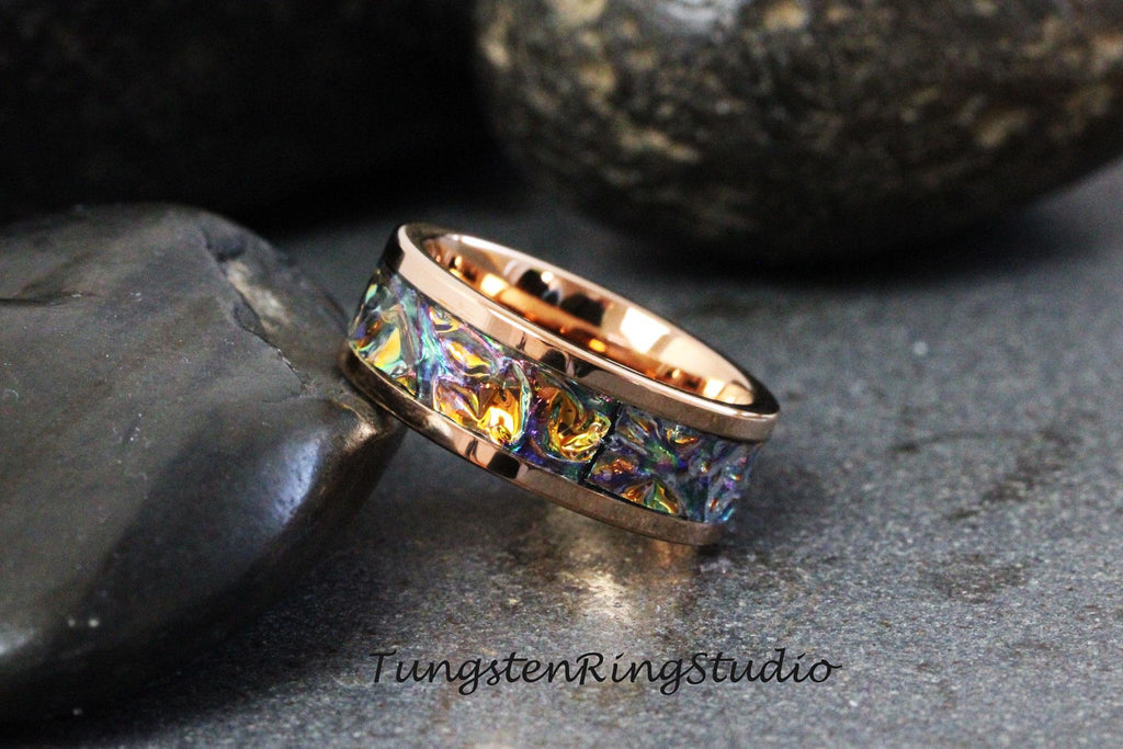 May include: A rose gold tungsten carbide ring with a colorful inlay of iridescent blue, green, and gold. The ring is on a dark gray background.