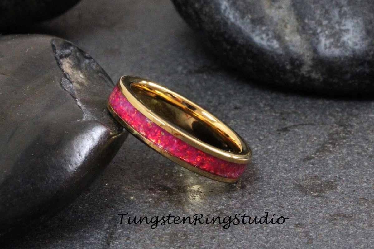 May include: A gold tungsten carbide ring with a pink opal inlay. The ring is on a dark gray background.